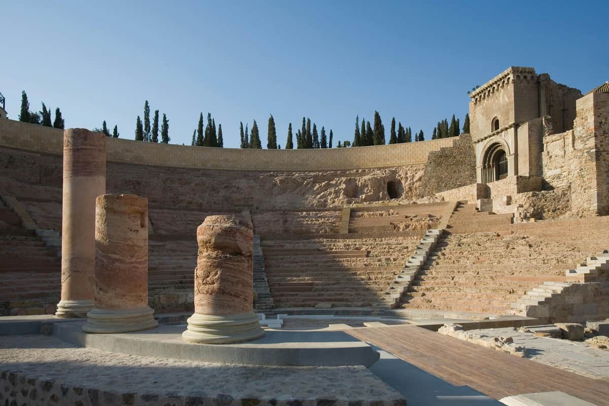 Museum of the Roman Theatre of Cartagena: Entry Ticket Ancient amphitheater with stone steps, columns in the foreground, and cypress trees on the horizon. Clear blue sky.