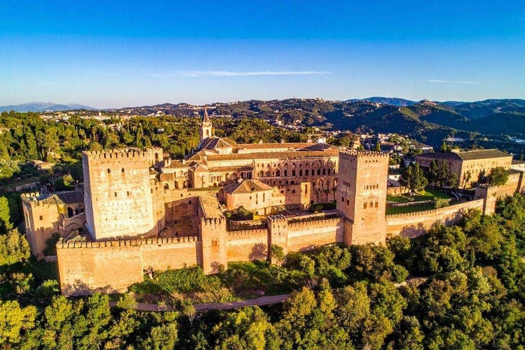 Alhambra: Guided Day Tour + City Pass A historic fortress complex on a hill surrounded by lush greenery, with a town and mountains in the background under a clear sky.