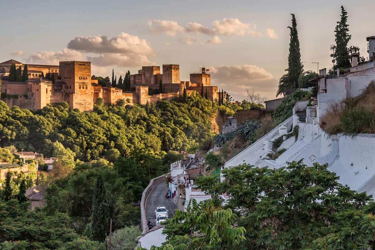 Alhambra, Nasrid Palaces, Albaicín & Sacromonte: Guided Tour Stone fortress with tall towers on a hilltop surrounded by lush greenery. A small road with white houses and parked cars below.