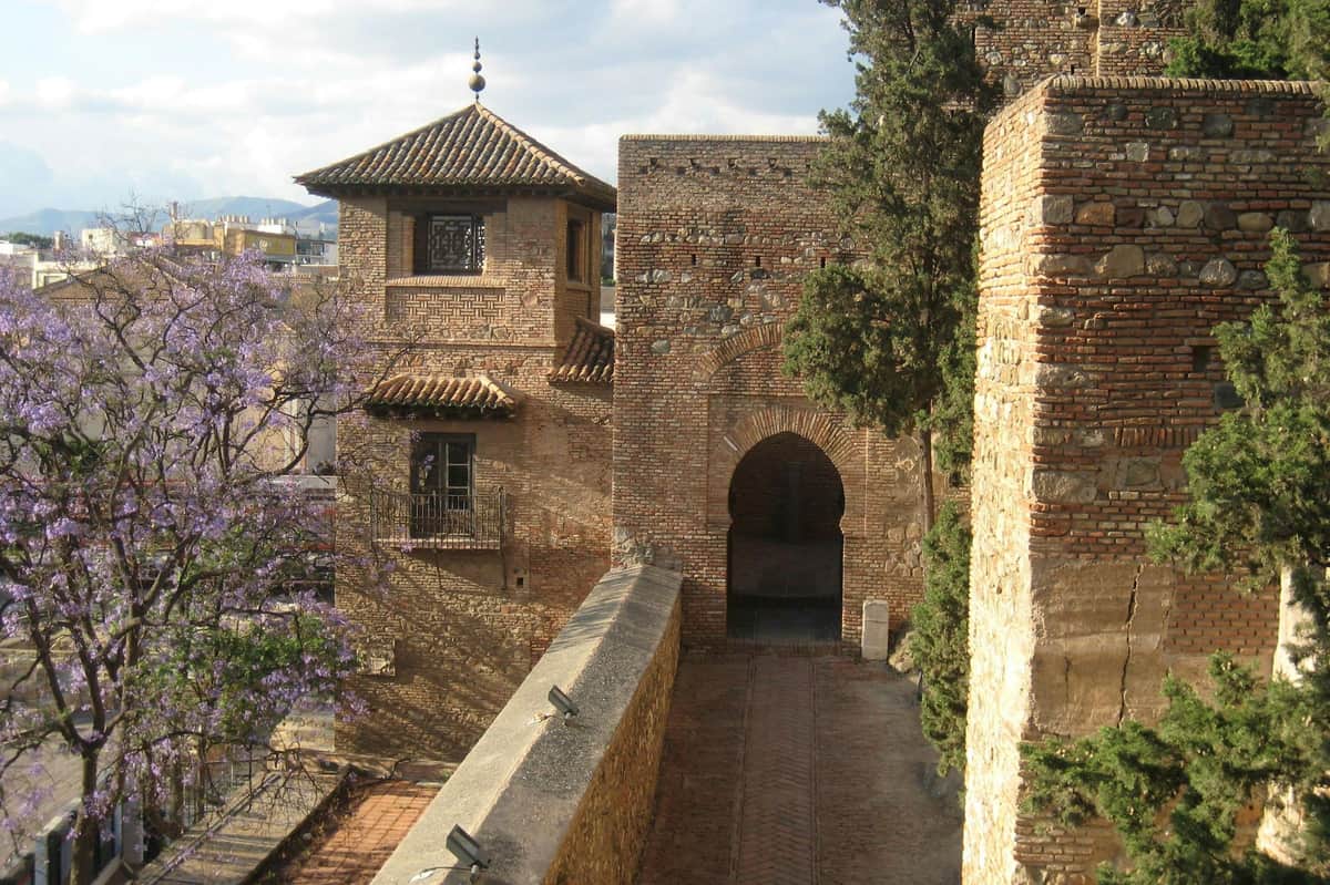 Málaga: Guided Walking Tour + Alcazaba Entry Ancient brick buildings with arched doorways and tiled rooftops, partially obscured by trees, under a partly cloudy sky.