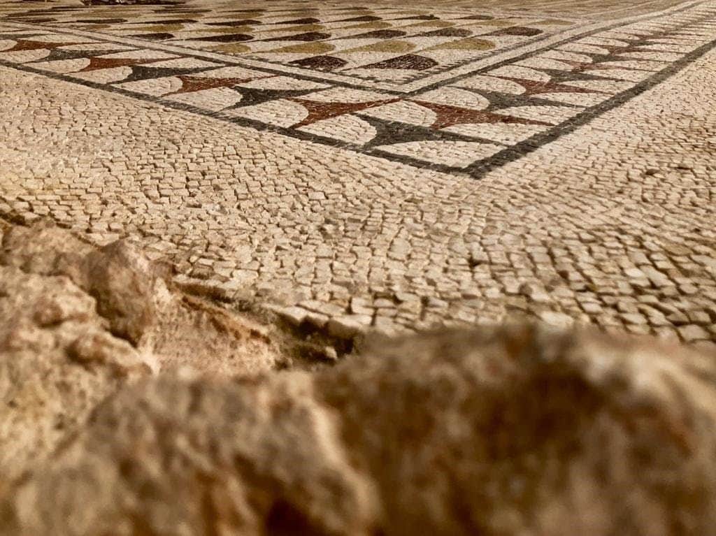 Roman Villa of Fuente Álamo Archaeological Site: Audio Guided Tour Close-up of an ancient mosaic tile floor with intricate geometric patterns, featuring a combination of cream, brown, and gray tiles.