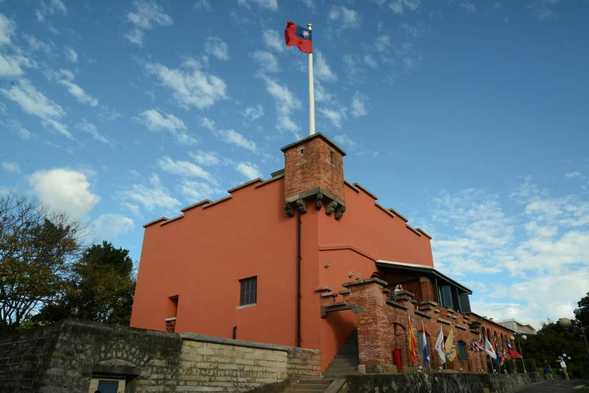 A red brick fort with a flagpole displaying a red flag under a blue sky with clouds.