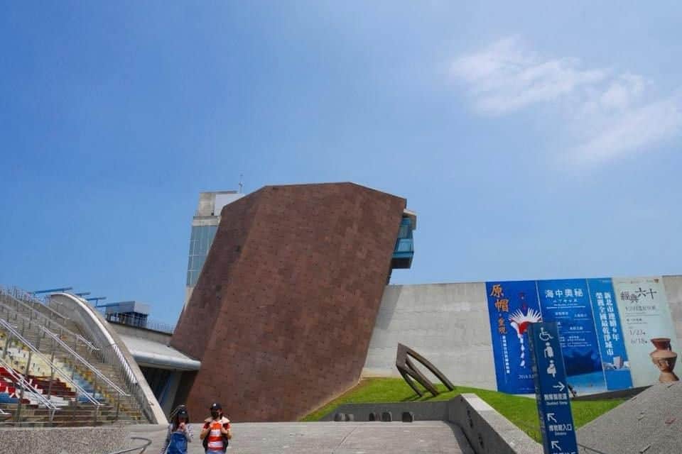 Musée archéologique de Shihsanhang à New Taipei : billet d'entrée + coffret « Thé de l'après-midi aux trésors culturels » Un bâtiment moderne aux lignes angulaires de couleur marron, flanqué de murs blancs, sous un ciel bleu limpide. Deux personnes marchent au premier plan.