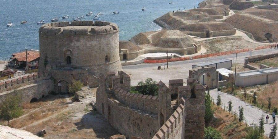 Troy: Day Trip with Lunch from Istanbul Stone fortress with round tower and crenelated walls overlooking a coastline with several boats in the sea.