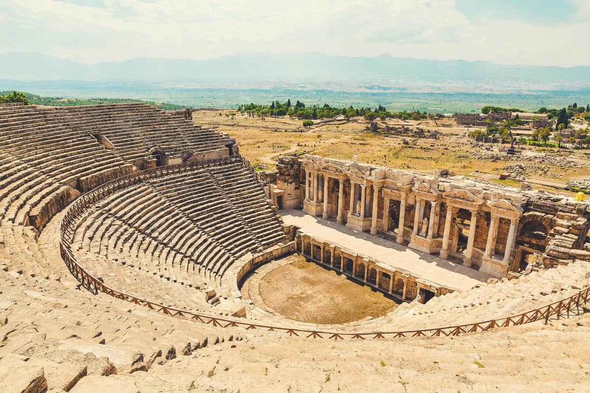Hierapolis & Travertines of Pamukkale: Day Tour from Antalya Ancient stone amphitheater with tiered seating and a columned stage, set in a vast, open landscape under a clear sky.