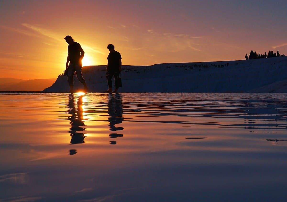 Pamukkale: Guided Tour Two people walking through shallow water during sunset, with a silhouetted landscape in the background.