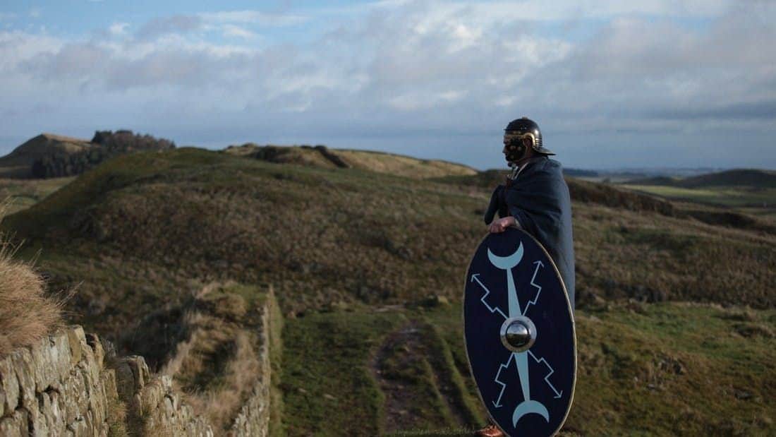 Hadrian's Wall: Half Day Tour From Newcastle A person wearing a helmet and cloak holds a shield while standing on a grassy hilltop under a cloudy sky.