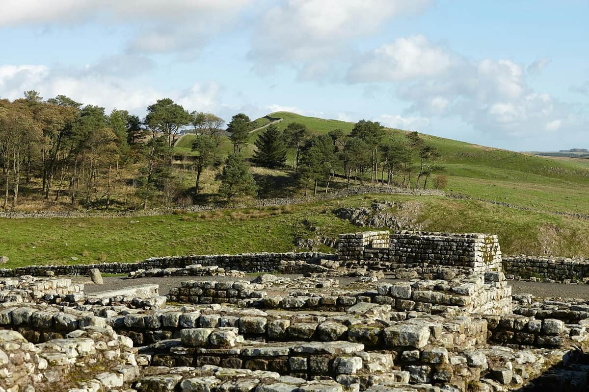 Housesteads Roman Fort – Hadrian's Wall Ancient stone ruins in the foreground with green rolling hills and trees under a partly cloudy sky in the background.