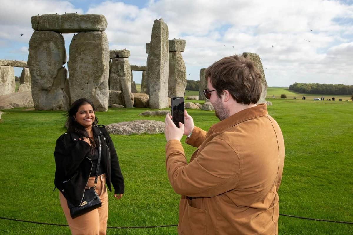 Stonehenge: Admission with Audio Guide + Premium Transport from London A person poses for a photo taken by another person in front of Stonehenge on a grassy field, under a partly cloudy sky.