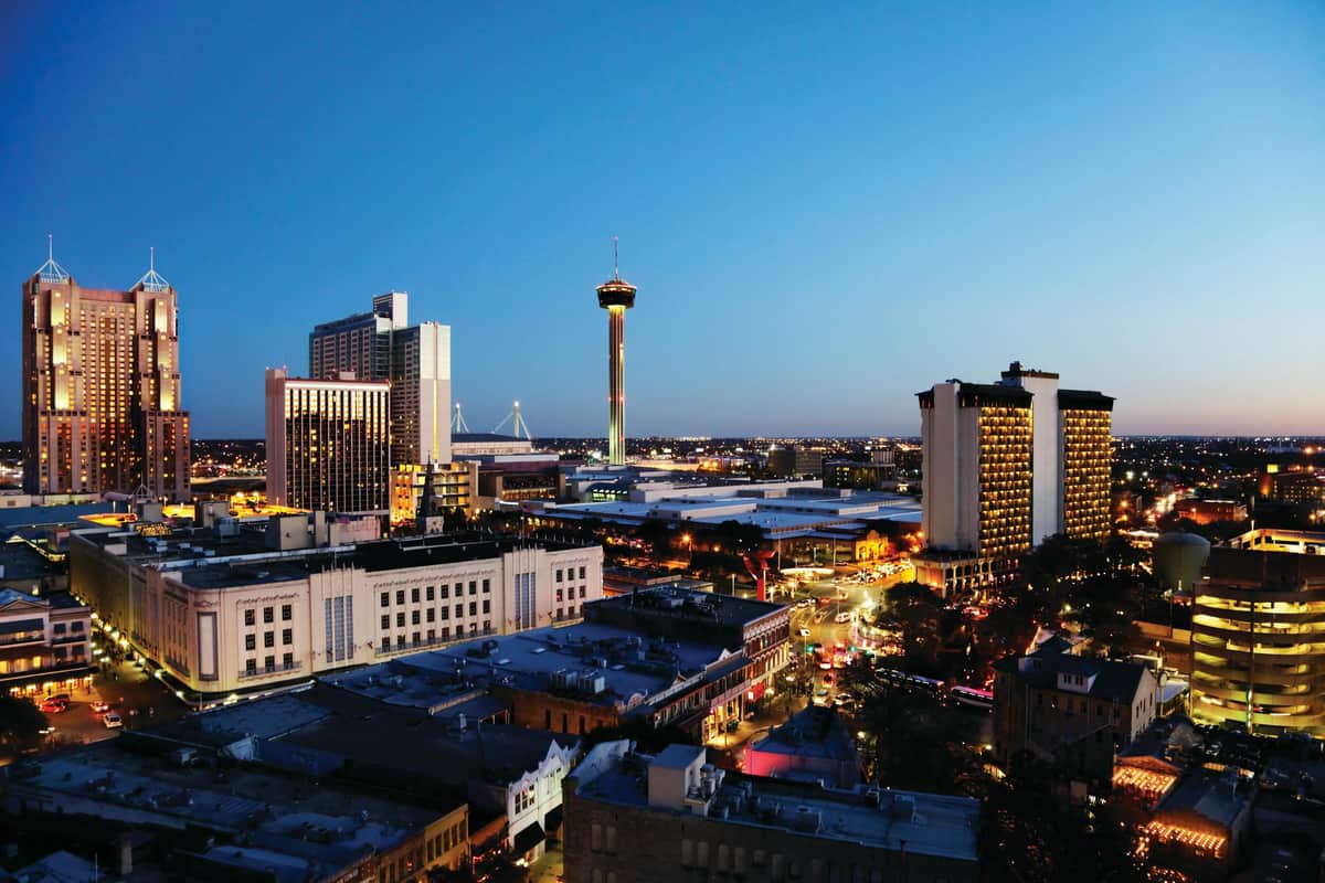 San Antonio: Highlights + Alamo Guided Tour Cityscape at dusk with tall buildings, a prominent tower in the center, and streets lit with car and building lights.