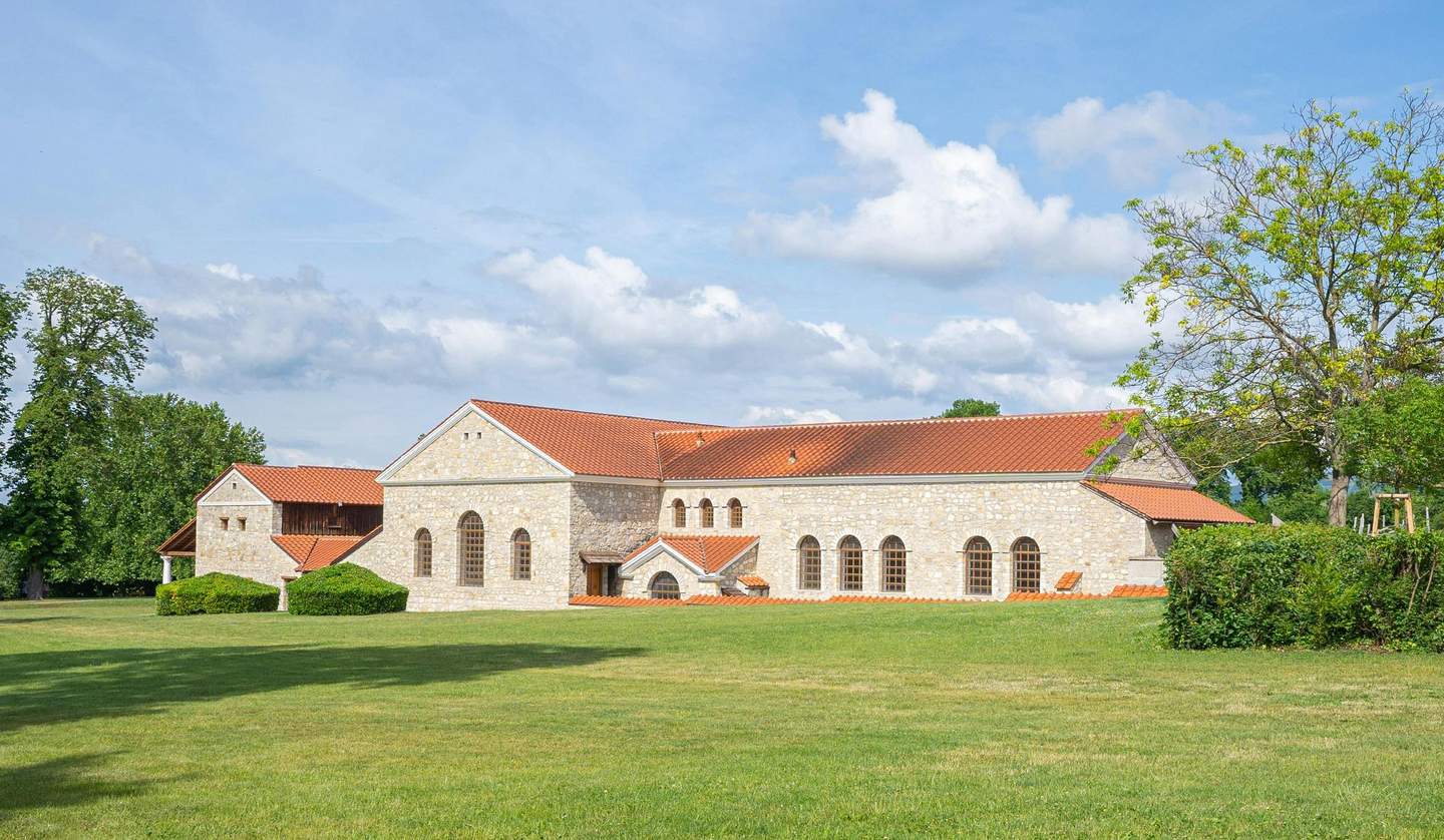 A stone building with arched windows and red-tiled roofs is surrounded by grassy lawns and trees under a blue sky.