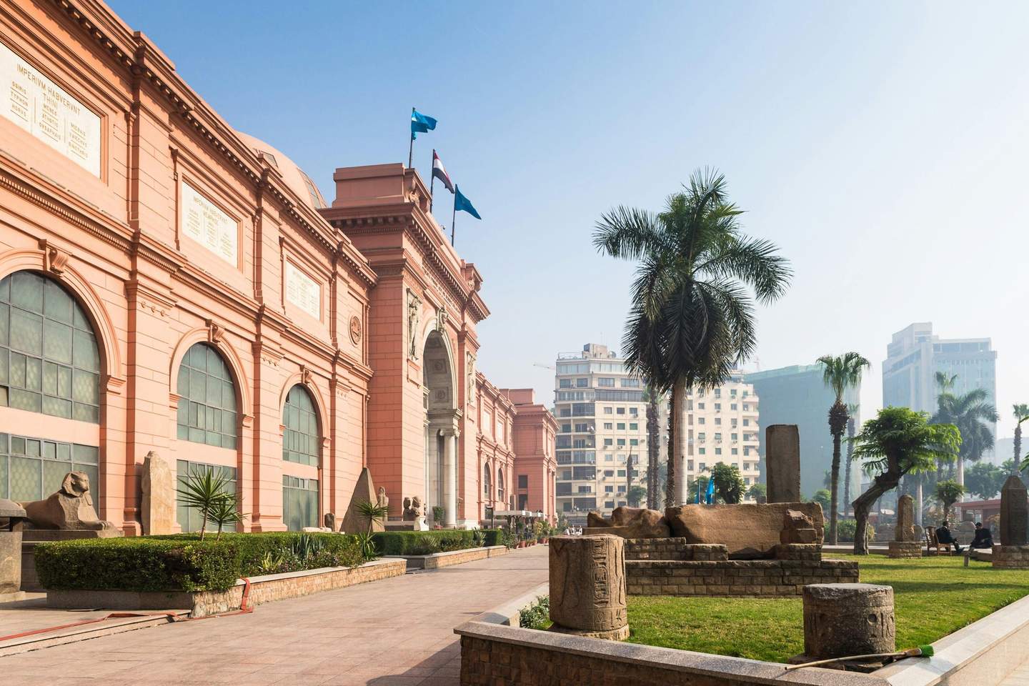 A large pink building with flags, architectural features, green plants, palm trees, and ancient artifacts in the foreground.