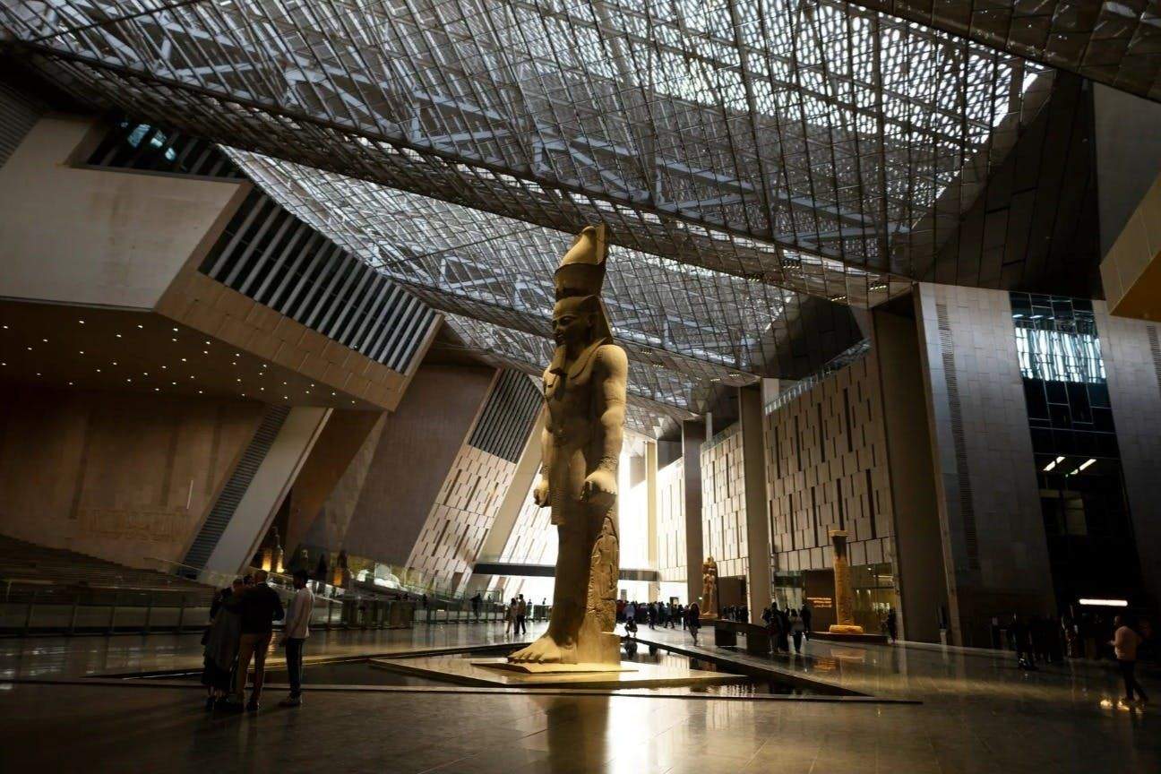 Large indoor hall with a giant statue of an ancient Egyptian pharaoh, modern architectural design, and small groups of people.