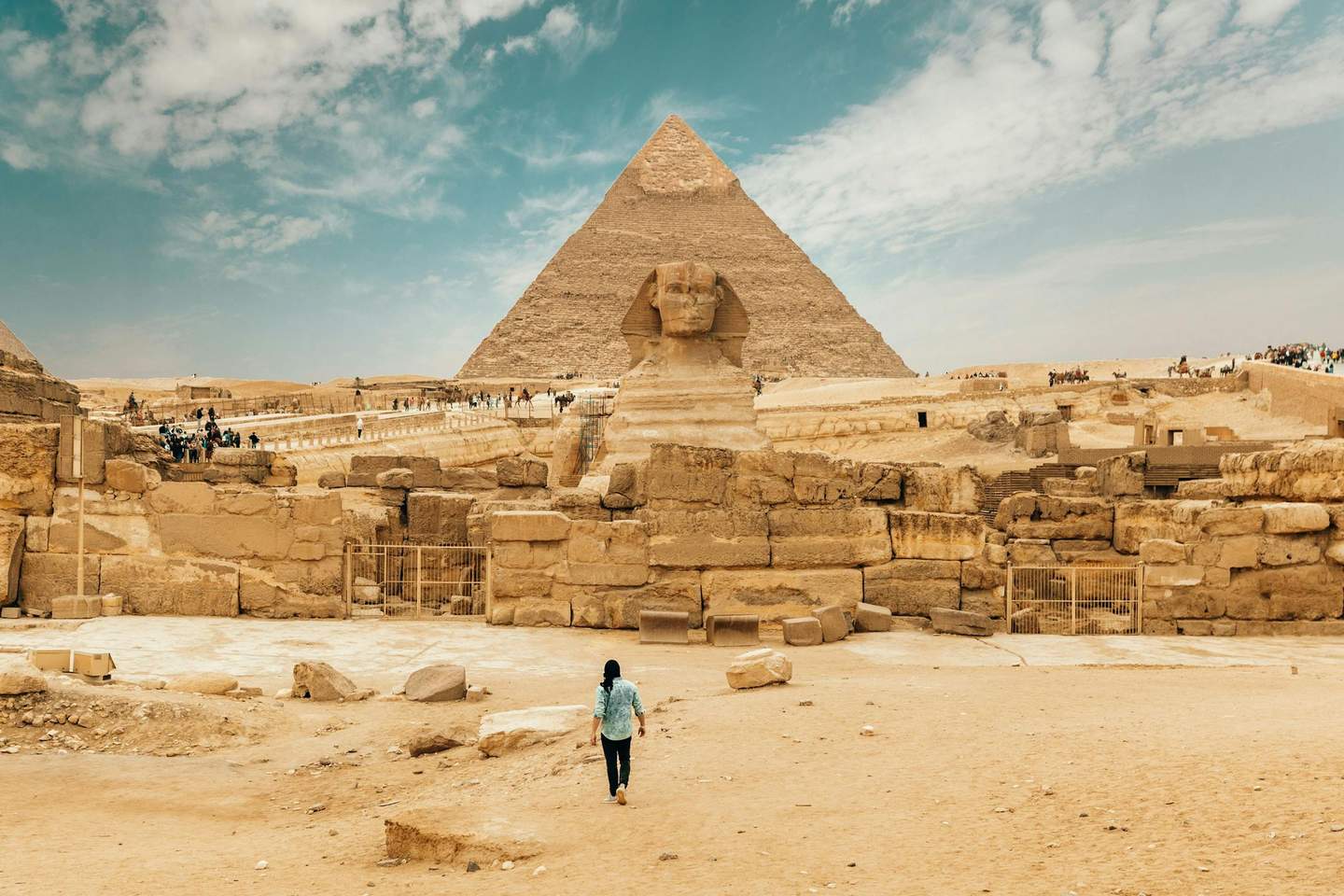 A person walks toward the Sphinx and a pyramid in Giza, with a desert landscape and scattered tourists in the background.