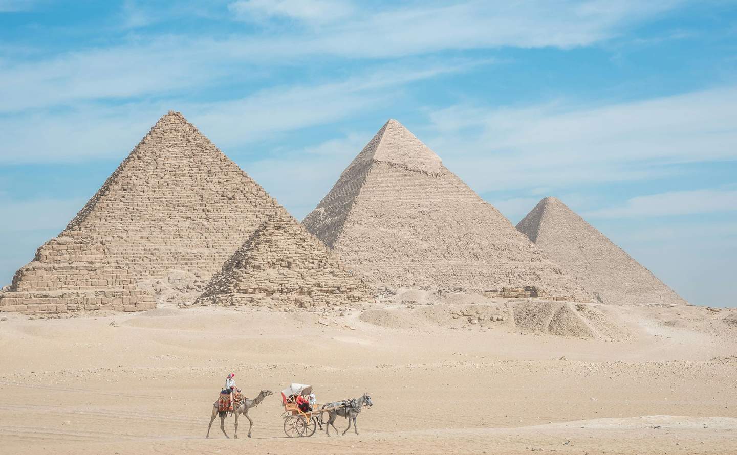 Three large pyramids in a desert with a clear blue sky, and a camel-drawn carriage transporting people in the foreground.