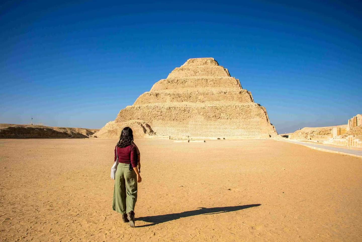 A person walking towards a step pyramid in a desert setting under a clear blue sky.