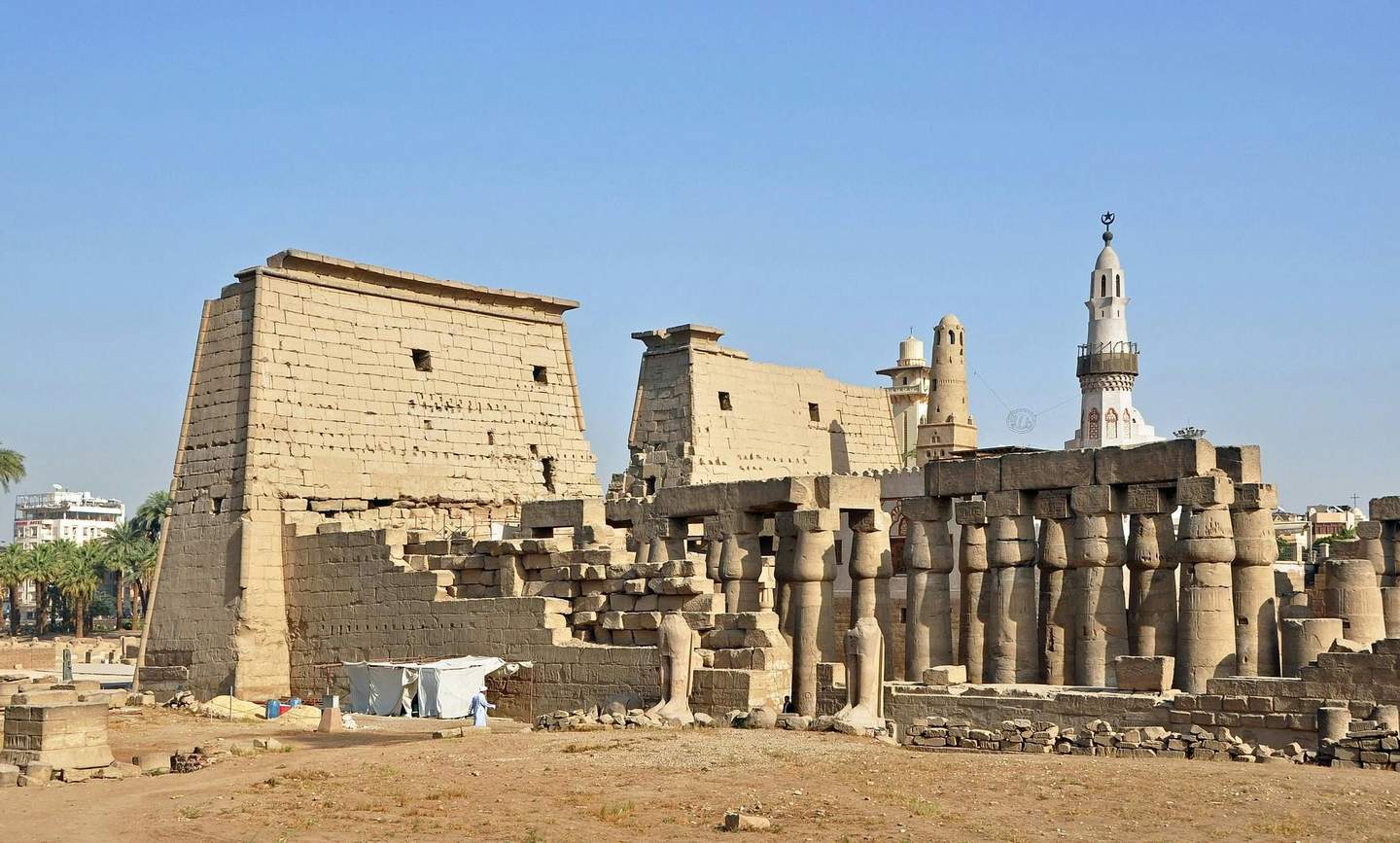 Ancient Egyptian temple ruins with large columns, a fragmented wall, and modern buildings in the background under a clear blue sky.