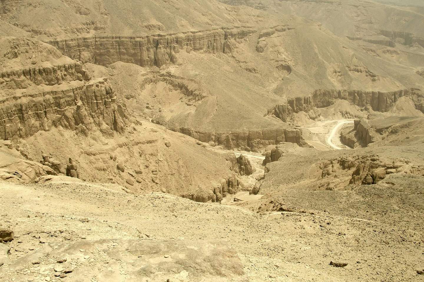 Arid desert canyon landscape with rugged terrain, exposed rock formations, and a winding dirt road in the valley.