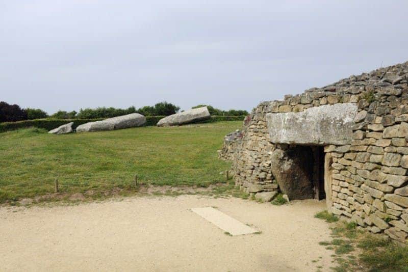Ancient stone structure with an entrance, surrounded by grass and two large horizontal stones in the background.