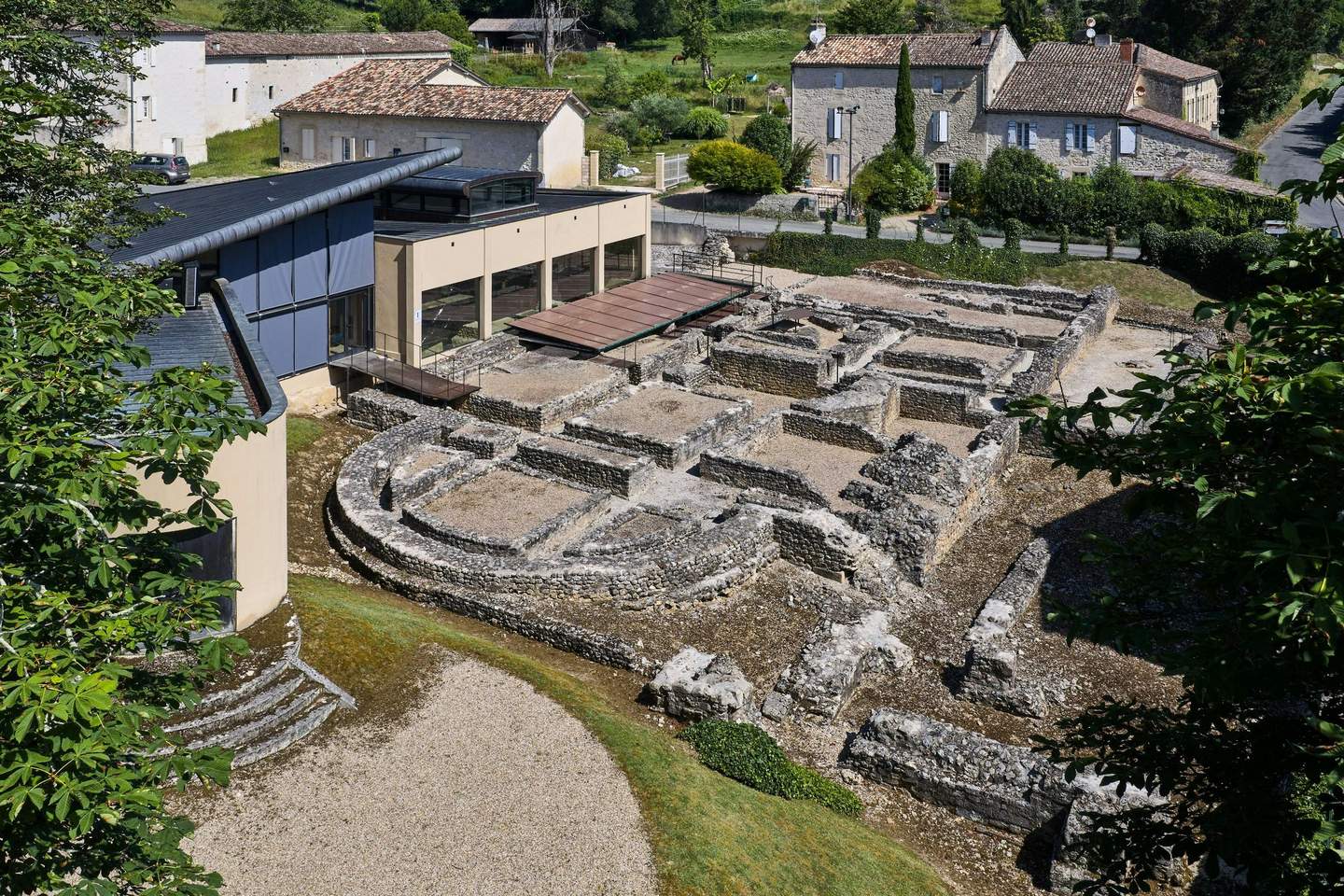 Ancient stone ruins beside a modern building with old houses and green vegetation in the background.