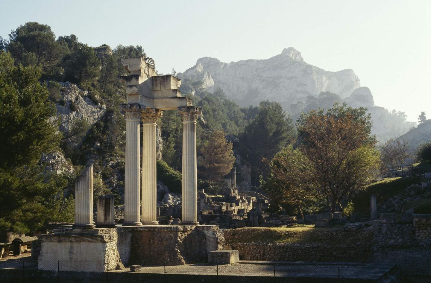 Ancient ruins with several tall columns amidst a hilly landscape with trees and distant mountains in the background.