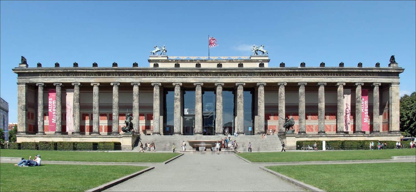 A neoclassical building with columns, two red banners reading "Altes Museum," and people walking and sitting on the steps and lawn.