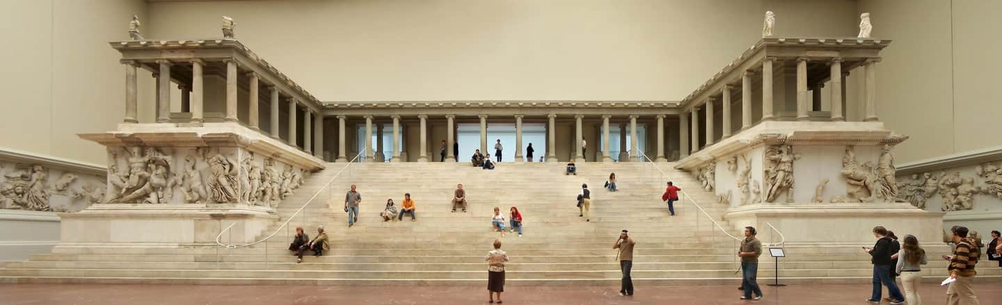 Pergamon Museum A grand stairway flanked by large architectural sculptures and columns, with people walking, sitting, and observing the artwork.