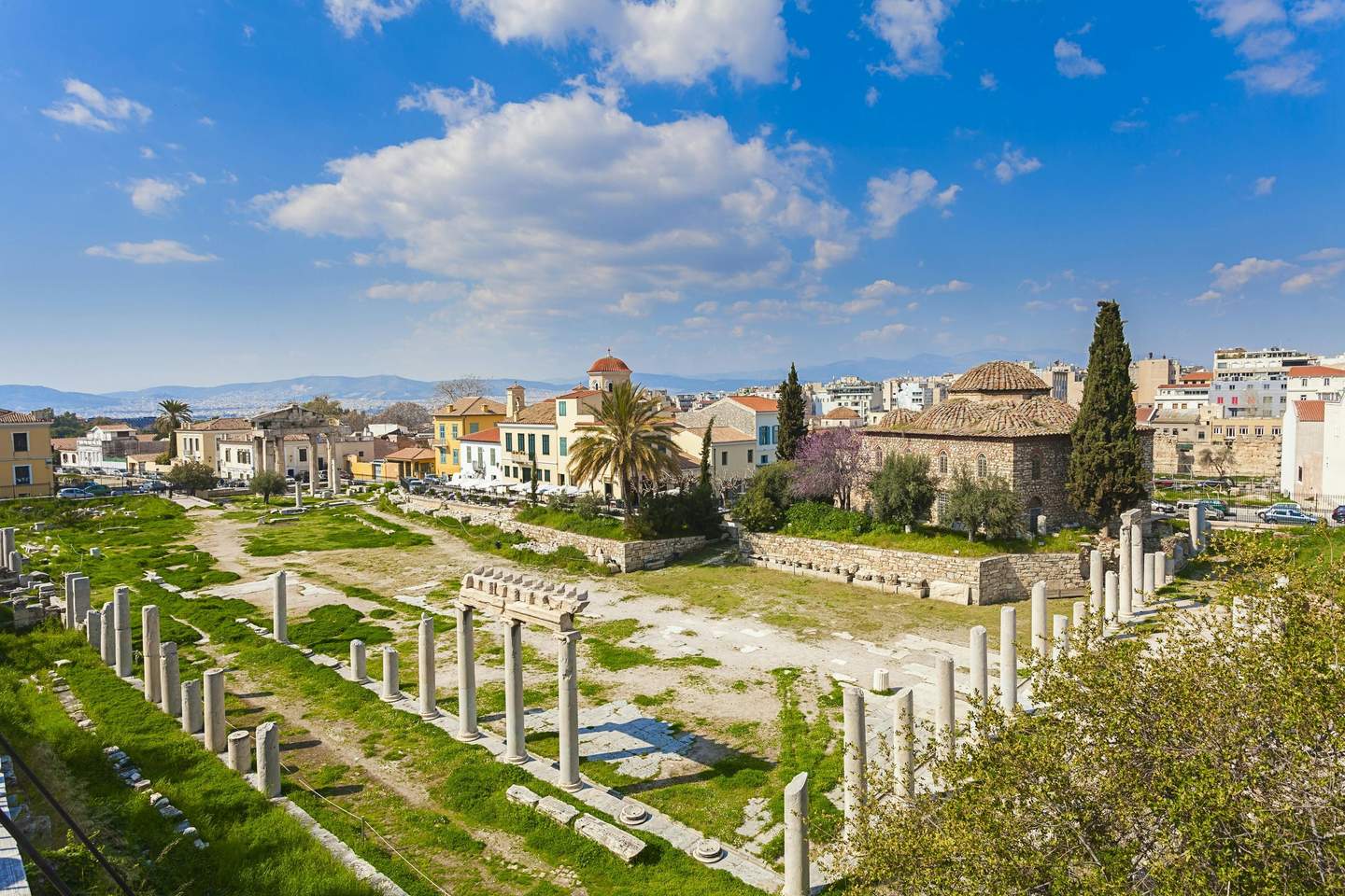 Ancient Agora of Athens Ancient ruins with stone columns and grassy areas, surrounded by historic buildings under a blue sky with scattered clouds.