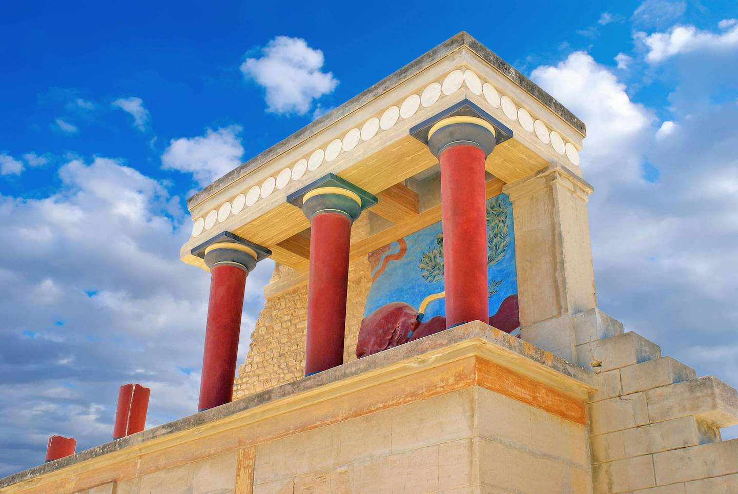 Palace of Knossos Ancient building with red columns, detailed mural, and a stepped structure, set against a blue sky with scattered clouds.
