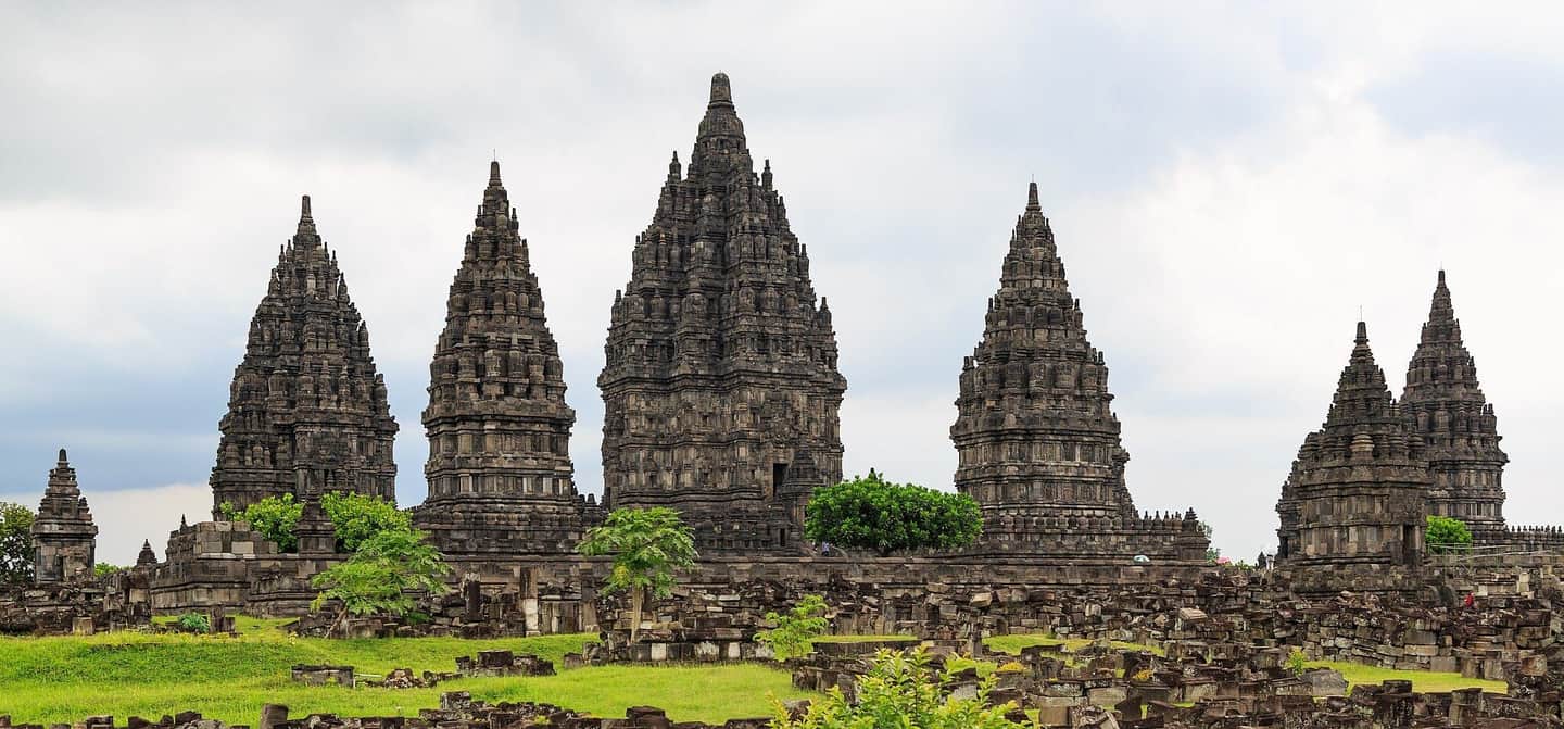 Several ancient, intricately carved stone temple towers rise above a grassy area with scattered ruins.