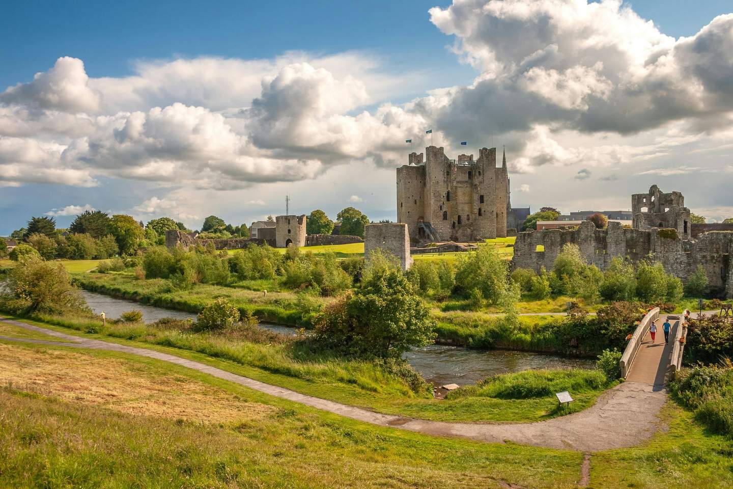 Trim Castle A medieval castle with tall towers, surrounded by ruins and lush greenery, a river, and a small bridge with people walking.