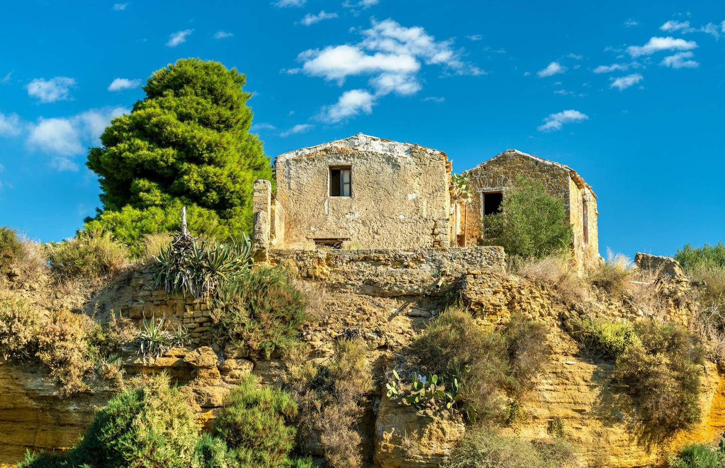 Kolymbethra Garden Two old stone buildings on a rocky hillside, surrounded by greenery and a large tree, under a blue sky with scattered clouds.