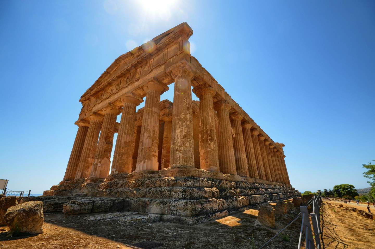 Ancient stone temple with columns under a bright blue sky, viewed from a low angle. Sparse trees and a dirt path are nearby.