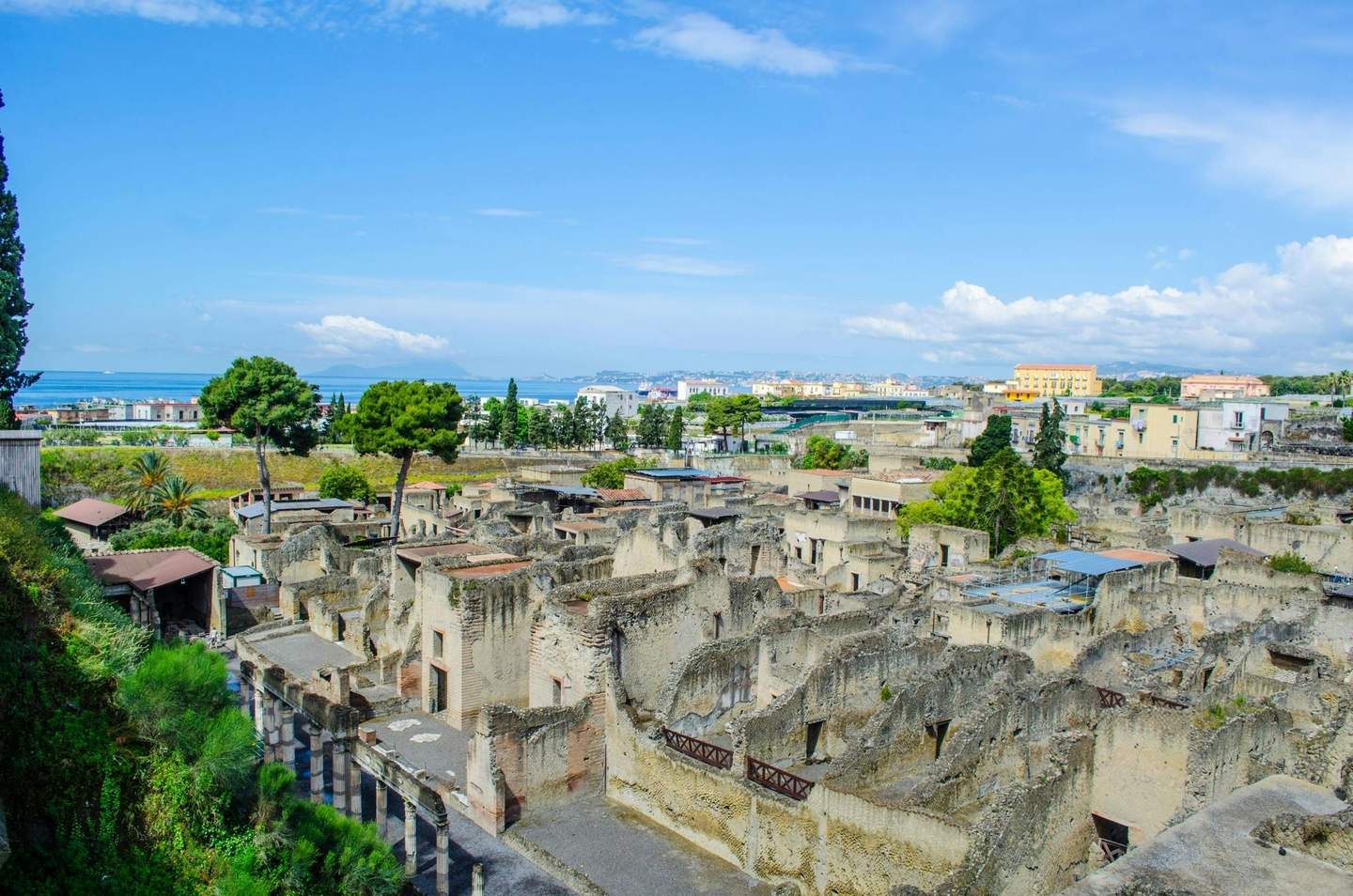 Ancient ruins with stone structures and columns, surrounded by greenery, with a town and sea in the background under a blue sky.