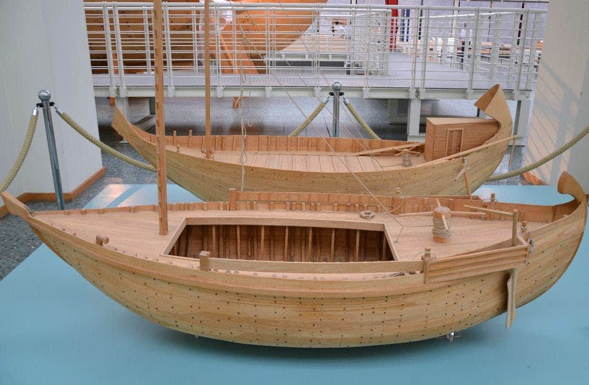 Two wooden boat models with detailed decks and masts on display, surrounded by railing barriers in an indoor setting.