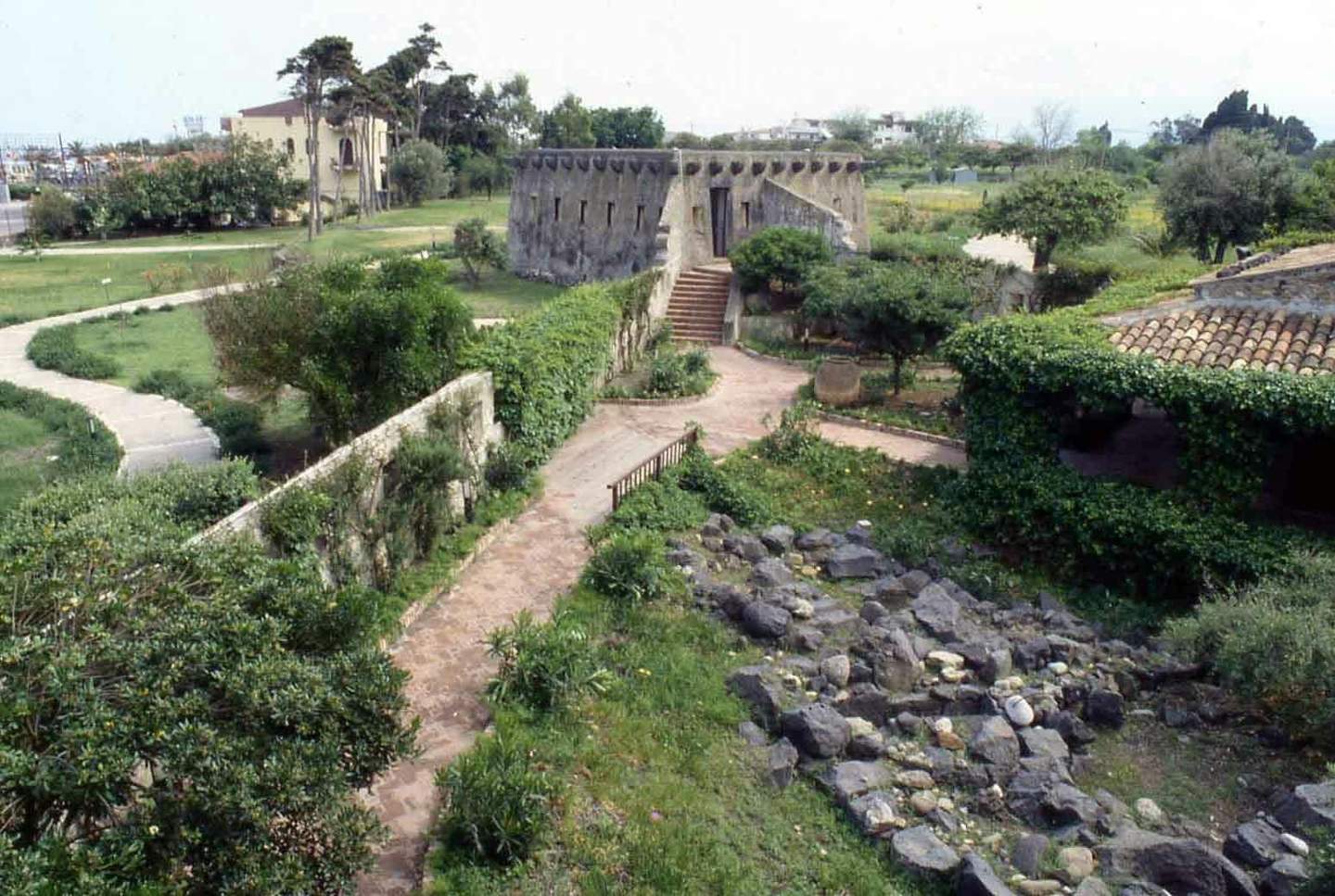 A historical building with stone walls is surrounded by pathways, greenery, trees, and scattered rocks in a park-like setting.