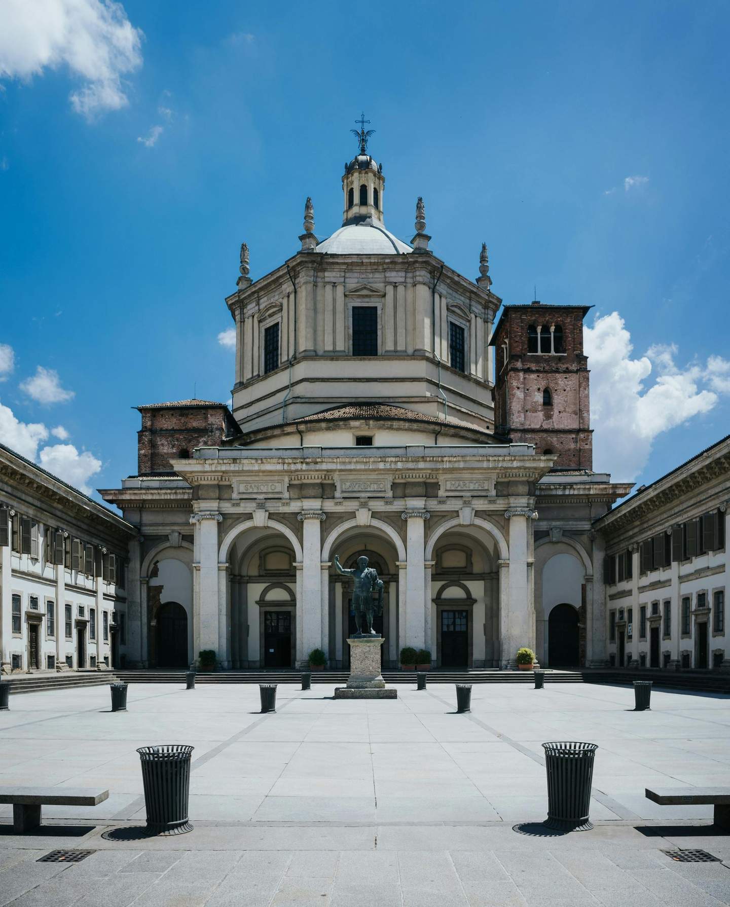 San Lorenzo Maggiore A historic building with a dome, arches, and a statue in the front courtyard, surrounded by columns and a clear blue sky.