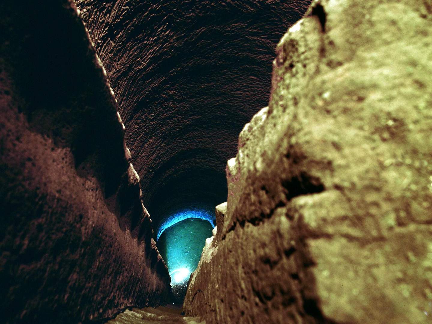 Pozzo della Cava A dimly lit, rocky tunnel with textured walls leading to a blue-lit area at the end.