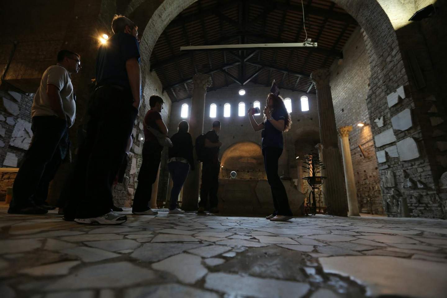 A group of people listen to a guide inside a historic stone building with high arches and columns.