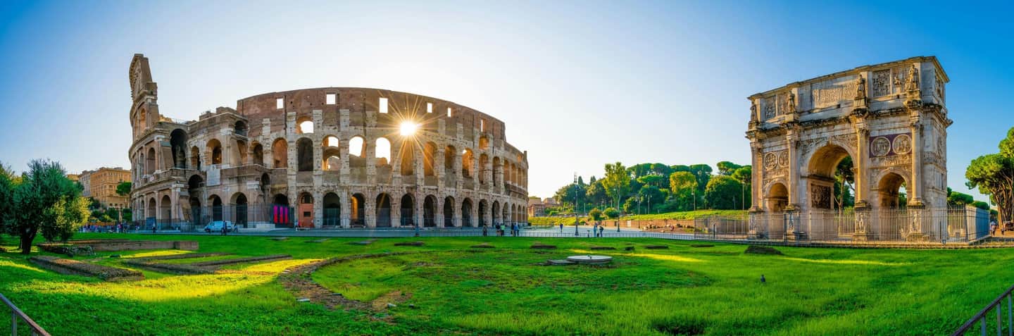 Ancient Colosseum and Arch of Constantine in Rome, with green grass and scattered visitors under a clear blue sky.