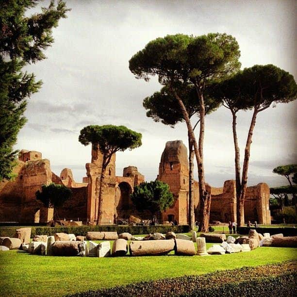 Tall trees surround ancient ruins and stone fragments on a green lawn under a cloudy sky.