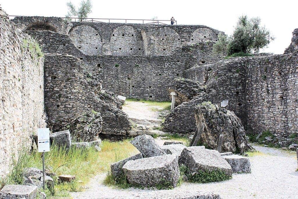 Ancient stone ruins with partially collapsed walls, scattered large stones, overgrown grass, and a person standing by a railing above.