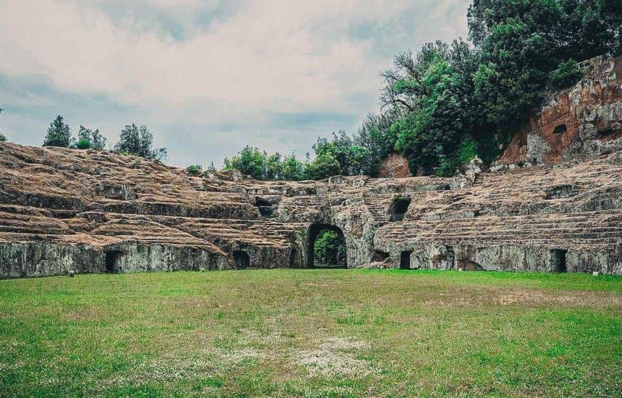 Amphitheater and Archaeological Park of Sutri Ancient stone amphitheater with tiered seating, grass center, and an arched entrance, surrounded by trees under a cloudy sky.