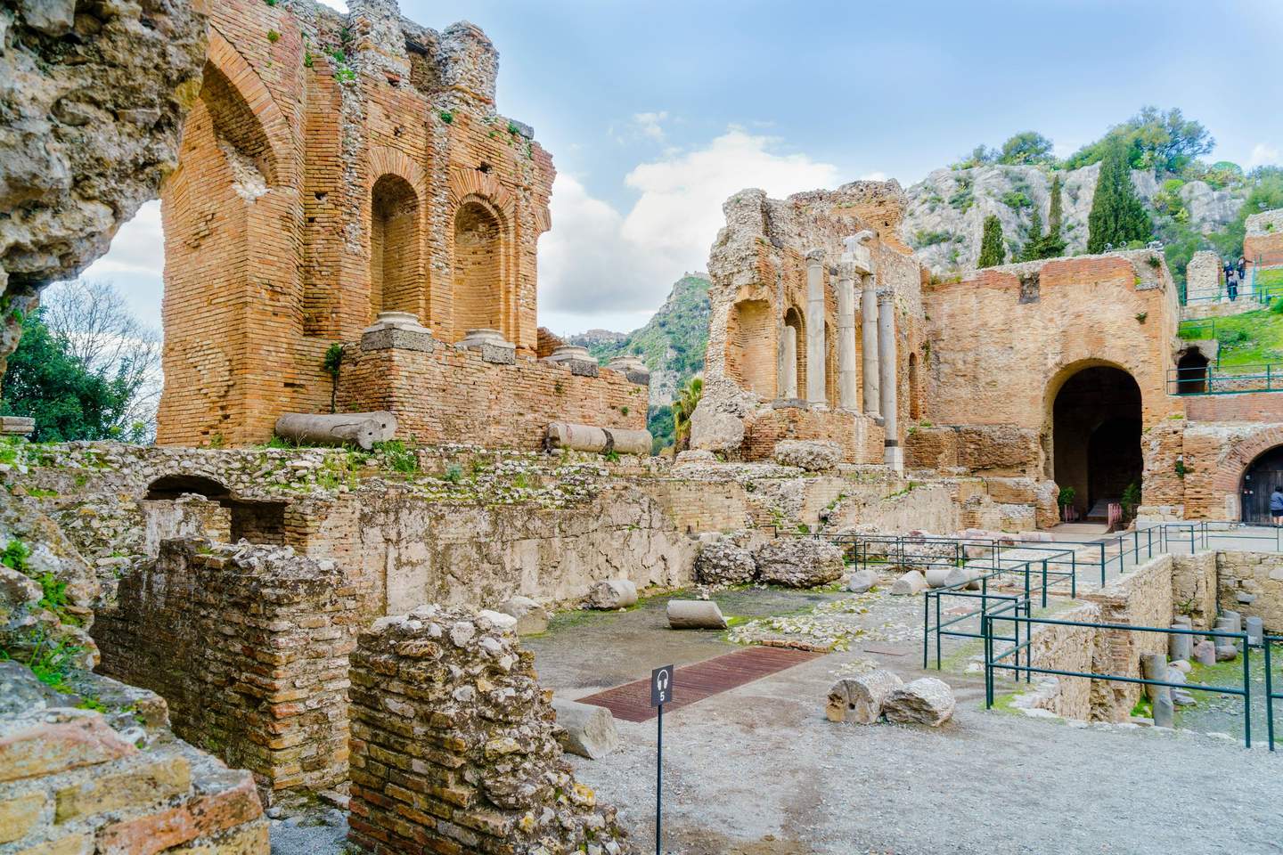 Ancient Theater of Taormina Ancient ruins with brick arches and columns in a rocky landscape with trees, pathways, and a cloudy sky.