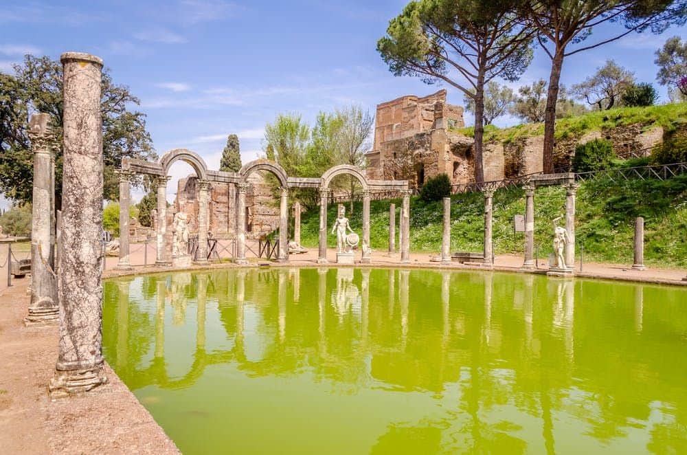Ancient ruins with statues and stone columns surround a green reflecting pool, set against a backdrop of trees and blue sky.