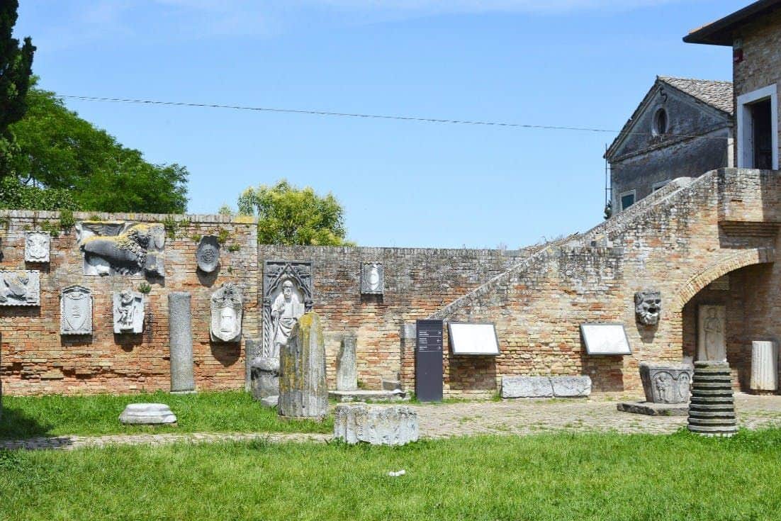 Museo Provinciale di Torcello A brick wall adorned with sculptures and plaques, surrounded by greenery, with a staircase and a building in the background.
