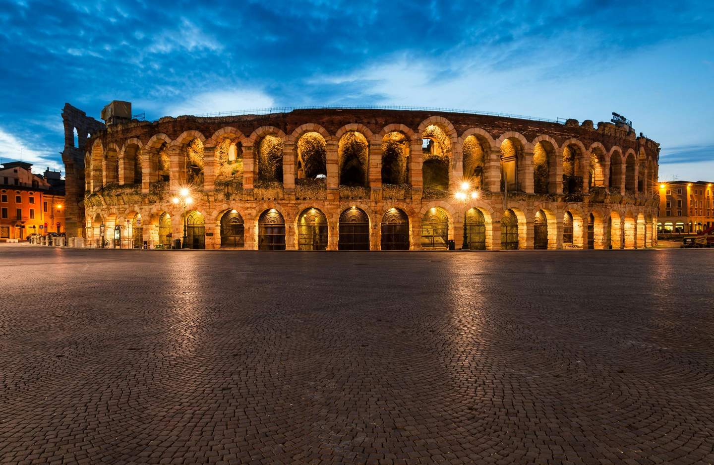 Ancient amphitheater with illuminated arches, standing on a cobblestone plaza under a twilight sky with a few lit buildings nearby.
