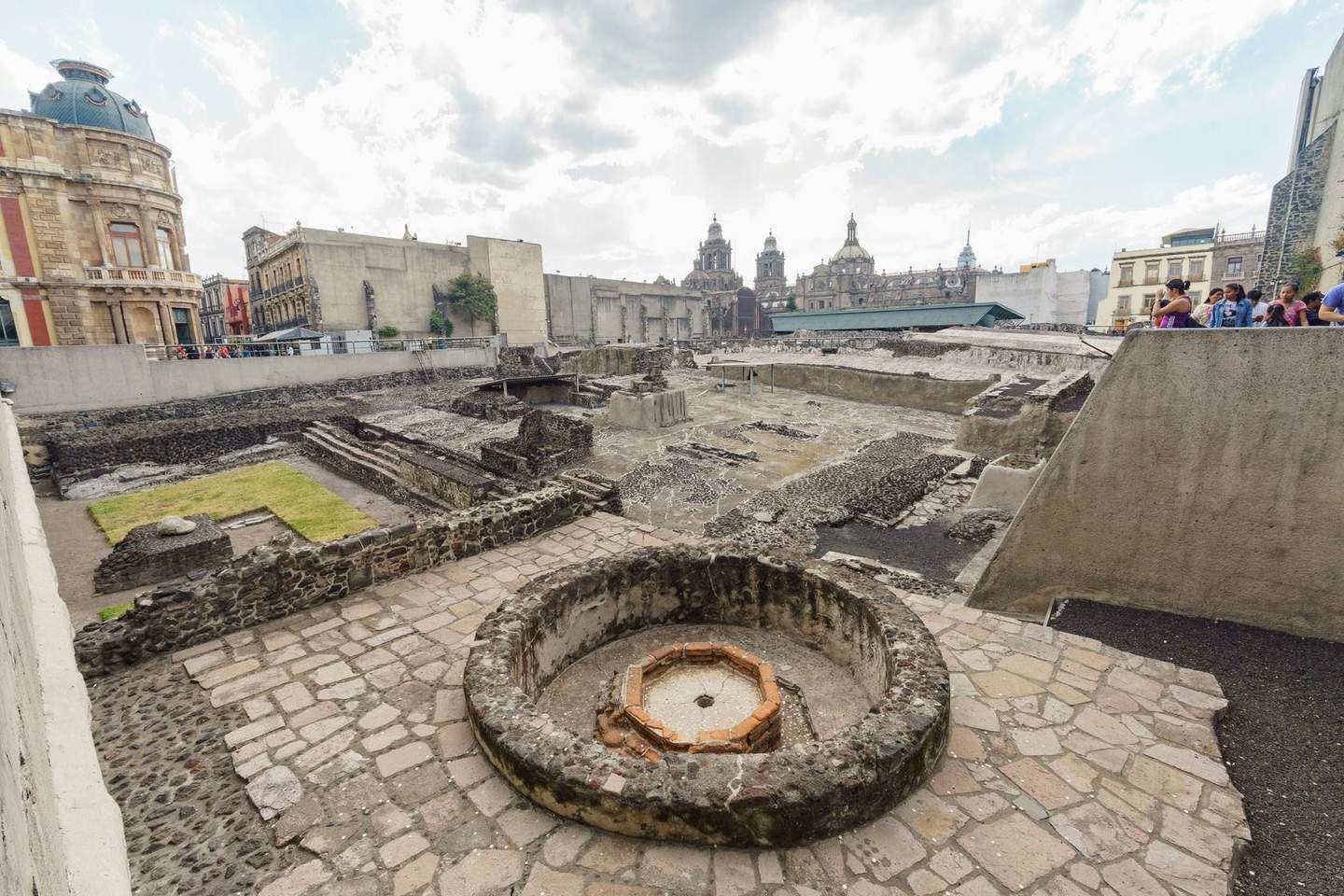 Templo Mayor Museum Ruins of ancient stone structures in an urban area with historic buildings in the background and a group of people observing.