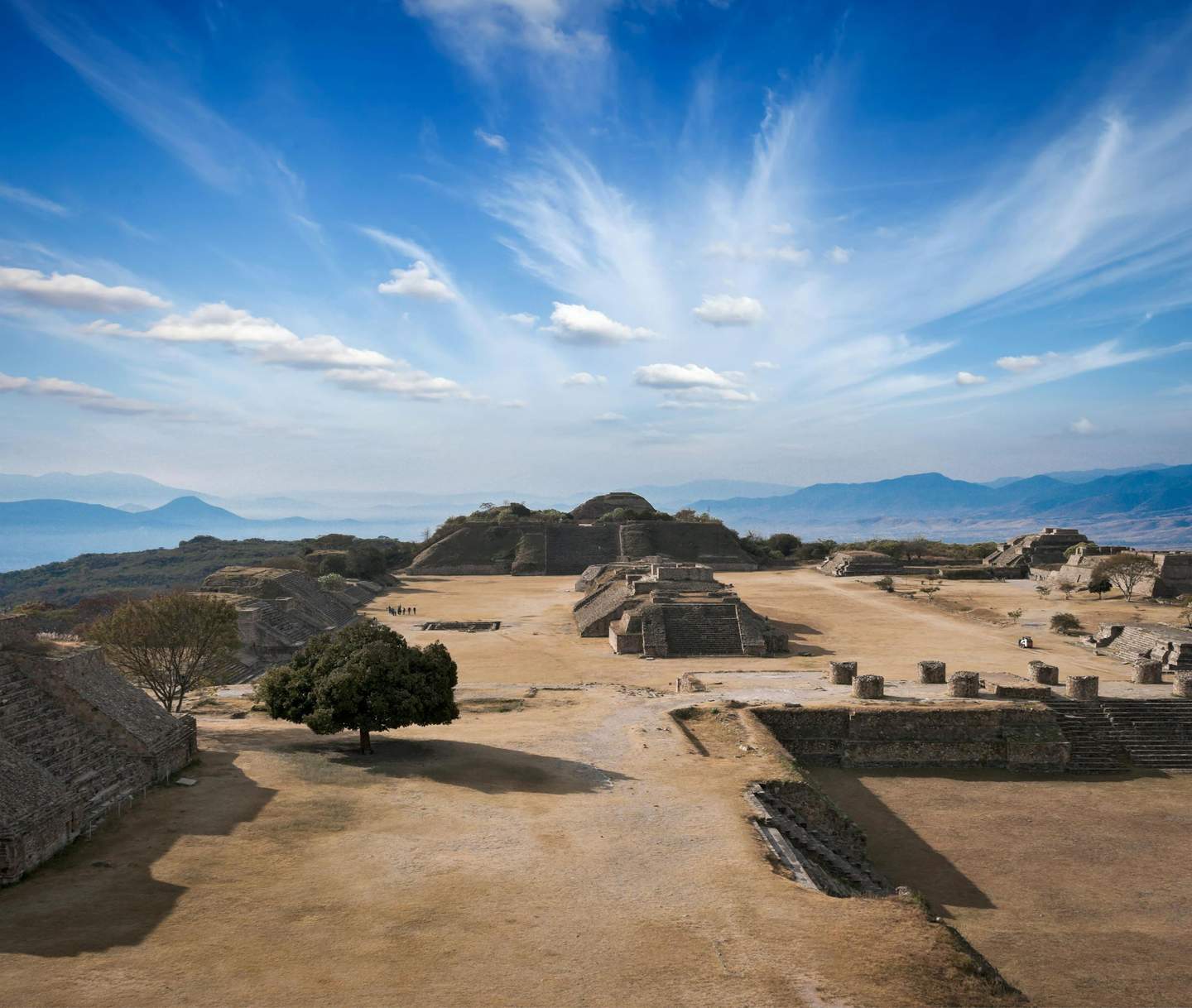 Monte Albán Archaeological Zone Ancient ruins on a plateau under a blue sky with scattered clouds, featuring stone structures and a lone tree.
