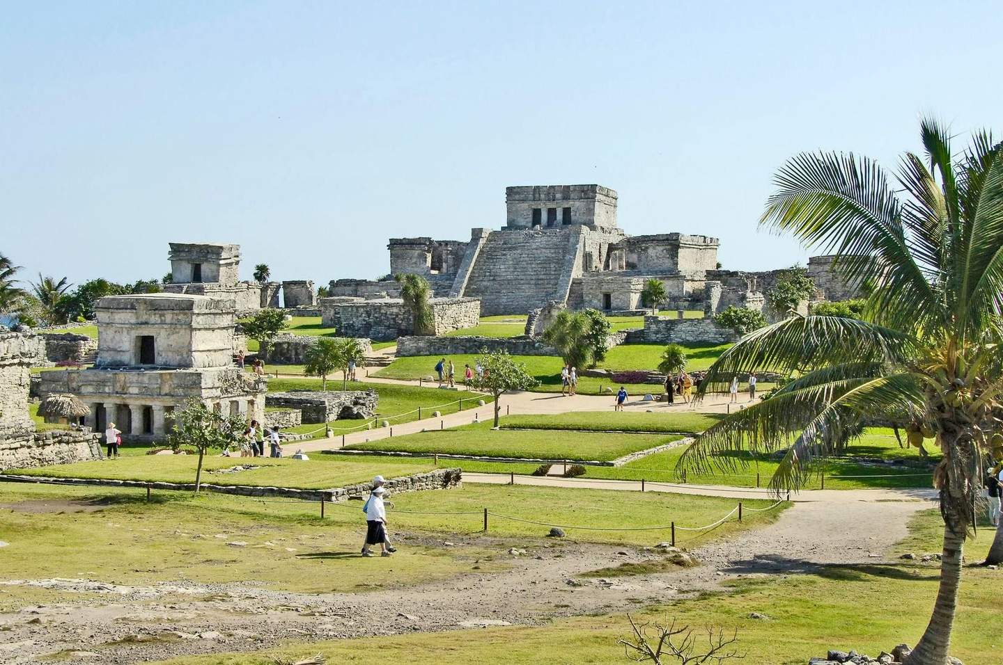 Mayan Ruins of Tulum Ancient stone ruins with pyramids and temples, visitors walking among the structures, and a palm tree in the foreground.